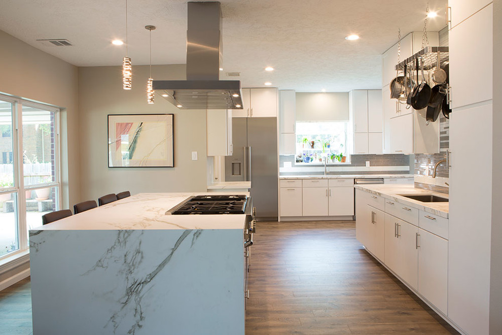 Expansive modern kitchen in Houston with a submerged sink, flat-panel cabinets, white cabinets, composite countertops, grey splashback, glass tiled splashback, stainless steel appliances, medium hardwood flooring and an island.