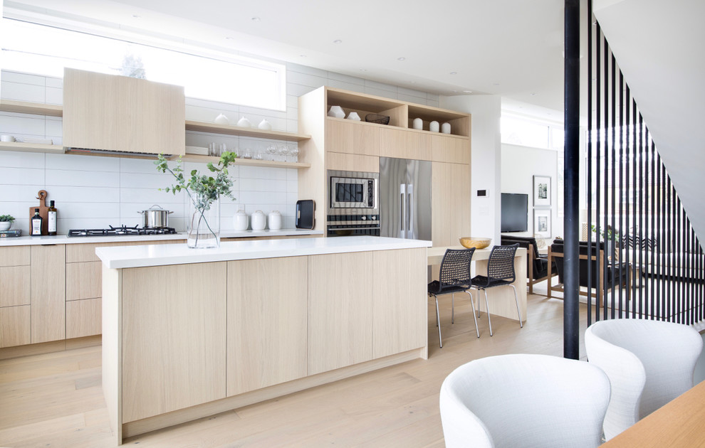 Photo of a scandi l-shaped kitchen/diner in Vancouver with flat-panel cabinets, light wood cabinets, white splashback, stainless steel appliances, light hardwood flooring, an island, beige floors and white worktops.