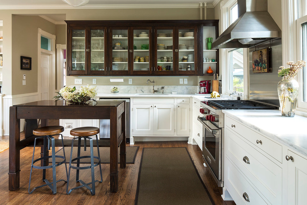 Exempel på ett amerikanskt l-kök, med en undermonterad diskho, luckor med glaspanel, skåp i mörkt trä, marmorbänkskiva, vitt stänkskydd, rostfria vitvaror, mörkt trägolv och en köksö