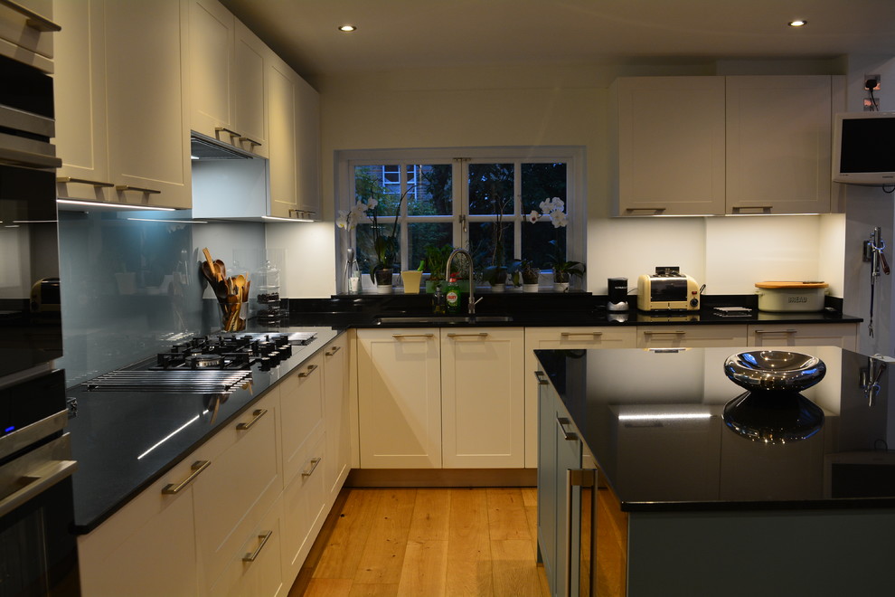 Пример оригинального дизайна: большая угловая кухня-гостиная в стиле кантри с фасадами в стиле шейкер, белыми фасадами, гранитной столешницей, синим фартуком, фартуком из стекла, светлым паркетным полом, островом, черной техникой и монолитной мойкой