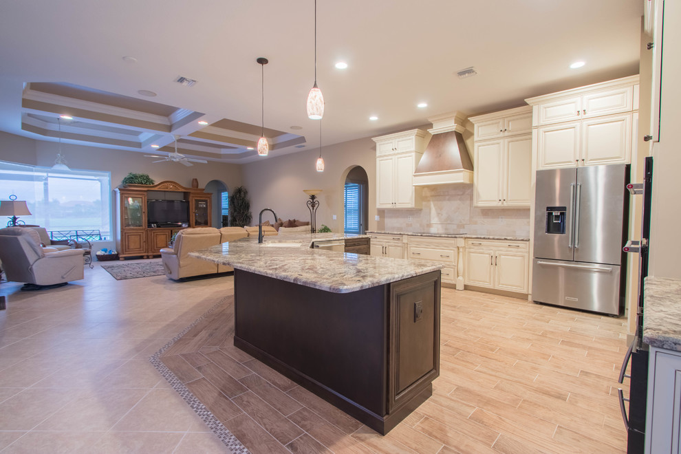 Example of a large classic l-shaped porcelain tile and brown floor open concept kitchen design in Other with a single-bowl sink, raised-panel cabinets, beige cabinets, granite countertops, beige backsplash, stone tile backsplash, stainless steel appliances and an island