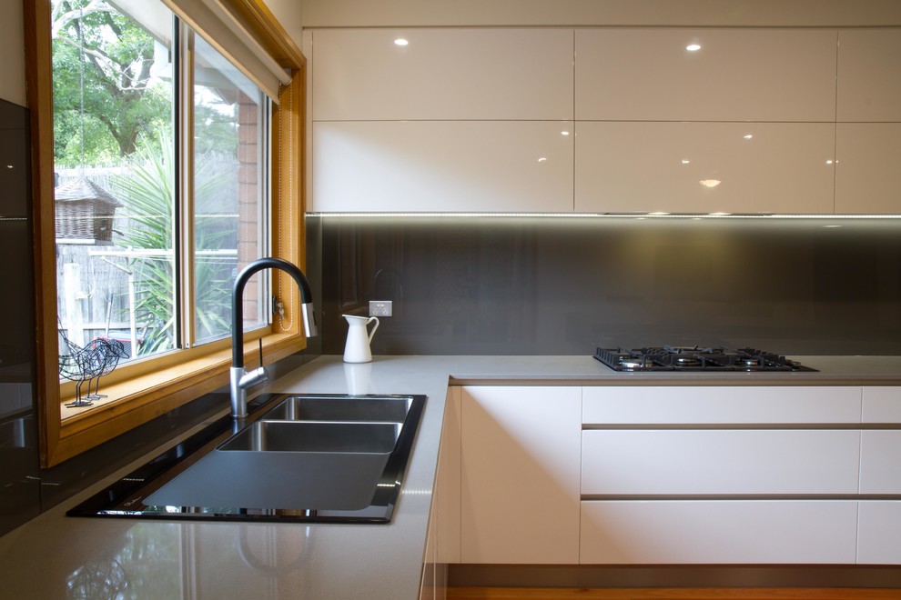 Cette image montre une grande cuisine américaine minimaliste en L avec un placard à porte plane, un plan de travail en quartz modifié, une crédence métallisée, une crédence en feuille de verre, un sol en bois brun et îlot.