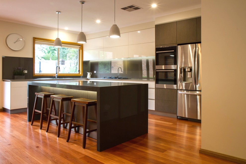 Große Moderne Wohnküche in L-Form mit flächenbündigen Schrankfronten, Quarzwerkstein-Arbeitsplatte, Küchenrückwand in Metallic, Glasrückwand, braunem Holzboden und Kücheninsel in Melbourne