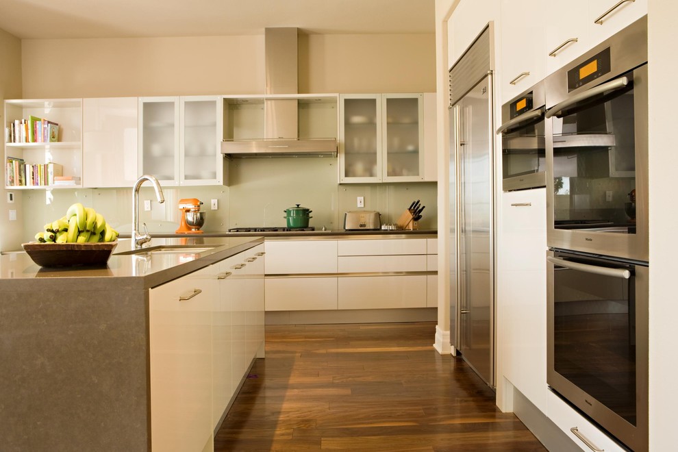 Moderne Küche mit Küchengeräten aus Edelstahl, Quarzwerkstein-Arbeitsplatte, Glasfronten, weißen Schränken und Glasrückwand in Los Angeles