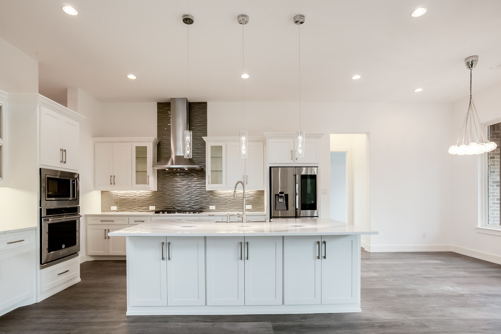 Photo of a large modern l-shaped open plan kitchen in Dallas with a belfast sink, recessed-panel cabinets, white cabinets, granite worktops, grey splashback, glass tiled splashback, stainless steel appliances, medium hardwood flooring, an island, brown floors and white worktops.