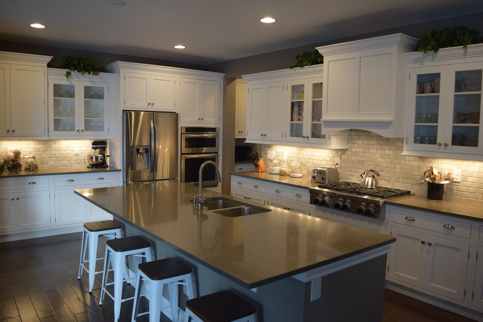 This is an example of a traditional l-shaped kitchen/diner in Denver with a submerged sink, flat-panel cabinets, white cabinets, an island, composite countertops, grey splashback, stone tiled splashback, stainless steel appliances and medium hardwood flooring.