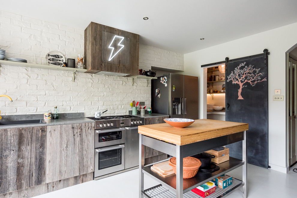 Medium sized eclectic single-wall kitchen in London with a built-in sink, flat-panel cabinets, concrete worktops, stainless steel appliances, concrete flooring, an island, grey worktops, distressed cabinets, white splashback, brick splashback and white floors.