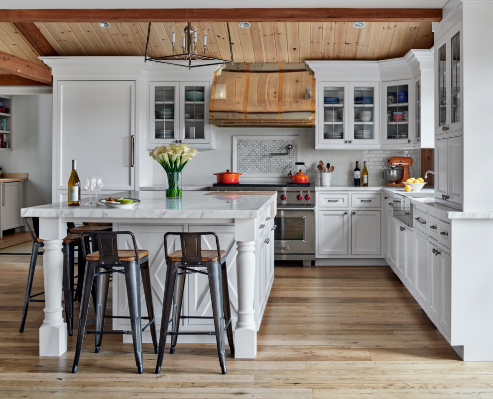 Photo of a country l-shaped kitchen in New York with white cabinets, white splashback, metro tiled splashback, stainless steel appliances, an island, beige floors and beaded cabinets.
