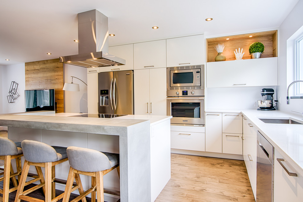 This is an example of a medium sized modern l-shaped kitchen/diner in Montreal with a submerged sink, flat-panel cabinets, white cabinets, concrete worktops, stainless steel appliances, an island, white splashback, medium hardwood flooring and brown floors.