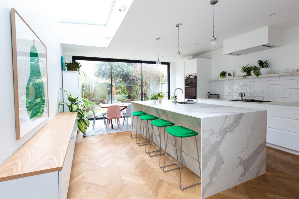 Large contemporary galley kitchen/diner in London with an integrated sink, flat-panel cabinets, white cabinets, marble worktops, white splashback, metro tiled splashback, light hardwood flooring, an island, white worktops, black appliances and brown floors.