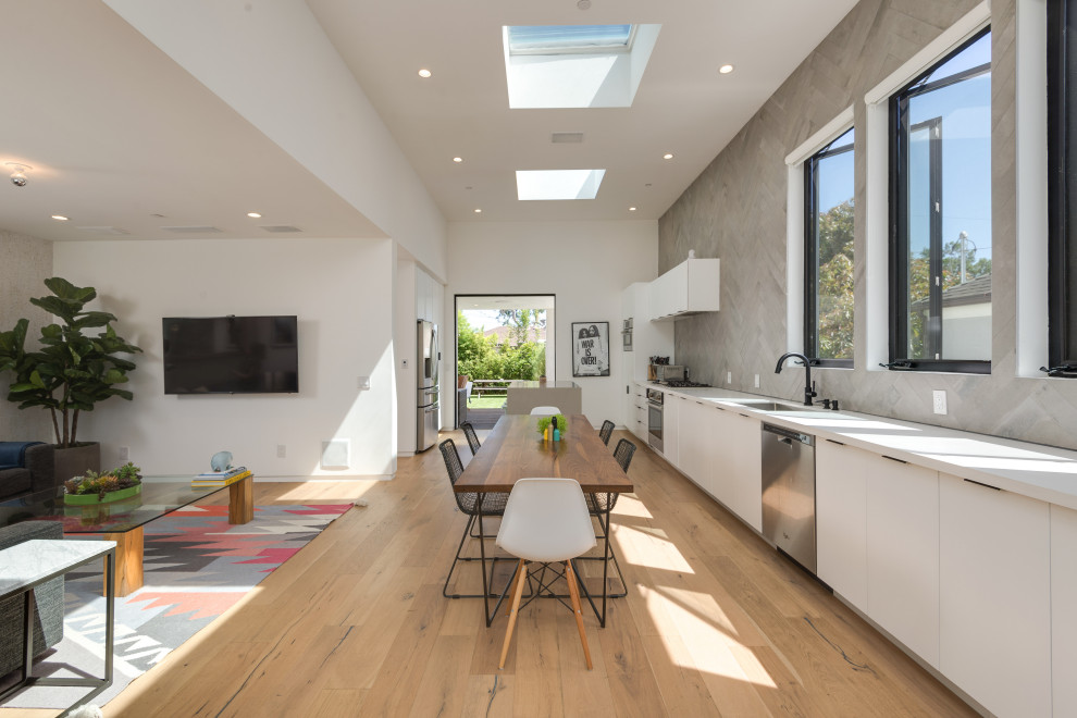 Inspiration for a medium sized contemporary single-wall open plan kitchen in Los Angeles with a single-bowl sink, flat-panel cabinets, white cabinets, engineered stone countertops, grey splashback, porcelain splashback, stainless steel appliances, white worktops, an island, medium hardwood flooring and brown floors.