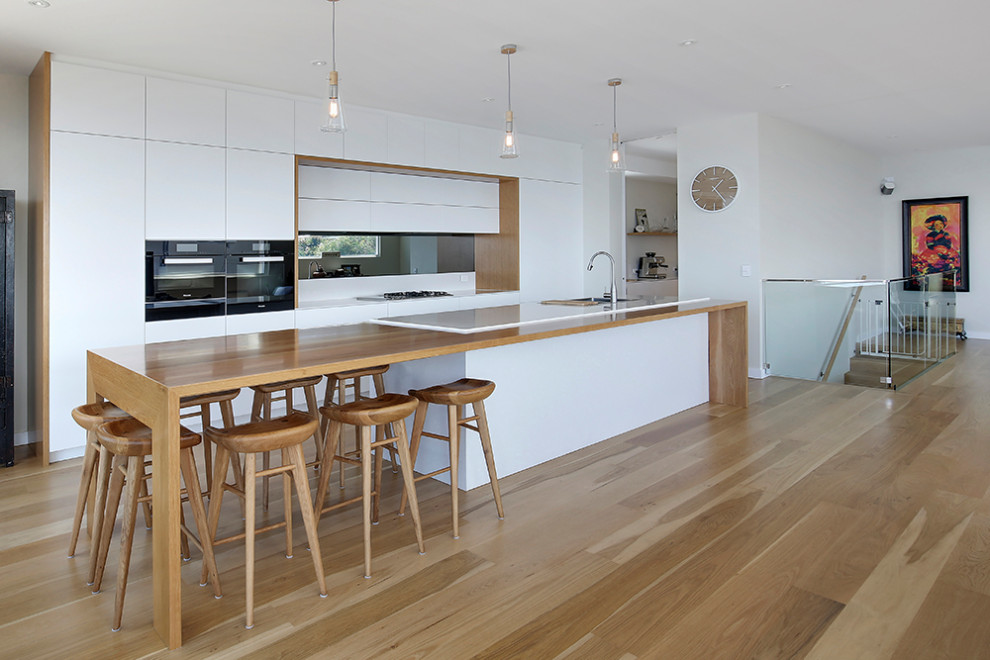 Inspiration for a large modern galley open plan kitchen in Sydney with a built-in sink, flat-panel cabinets, white cabinets, engineered stone countertops, metallic splashback, mirror splashback, stainless steel appliances, light hardwood flooring, an island, multi-coloured floors, white worktops and a coffered ceiling.