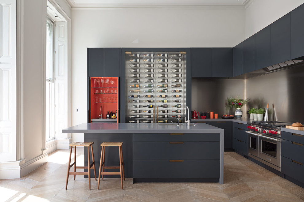 Inspiration for an expansive contemporary galley open plan kitchen in London with a submerged sink, glass-front cabinets, grey cabinets, composite countertops, light hardwood flooring, an island, brown floors, grey worktops, metallic splashback, metal splashback and stainless steel appliances.