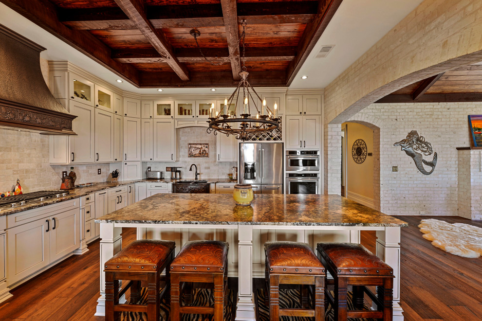 Maritim inredning av ett mellanstort brun brunt kök, med en rustik diskho, skåp i shakerstil, vita skåp, granitbänkskiva, beige stänkskydd, stänkskydd i travertin, rostfria vitvaror, mellanmörkt trägolv, en köksö och brunt golv