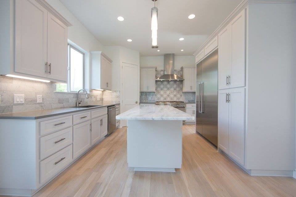 Photo of a medium sized contemporary l-shaped open plan kitchen in Austin with a submerged sink, recessed-panel cabinets, white cabinets, grey splashback, stone tiled splashback, stainless steel appliances, light hardwood flooring, an island and brown floors.