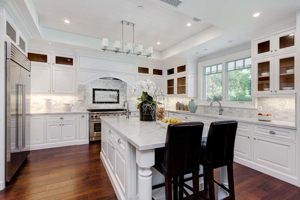 Klassisk inredning av ett stort kök, med en undermonterad diskho, luckor med upphöjd panel, vita skåp, marmorbänkskiva, vitt stänkskydd, stänkskydd i stenkakel, rostfria vitvaror, mellanmörkt trägolv och en köksö