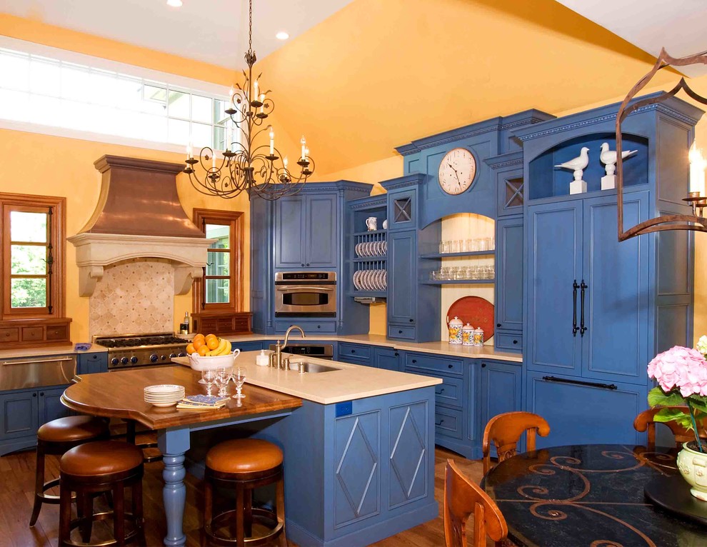 Example of a mid-sized country l-shaped medium tone wood floor enclosed kitchen design in Louisville with an undermount sink, recessed-panel cabinets, blue cabinets, solid surface countertops, beige backsplash, stone tile backsplash, stainless steel appliances and two islands