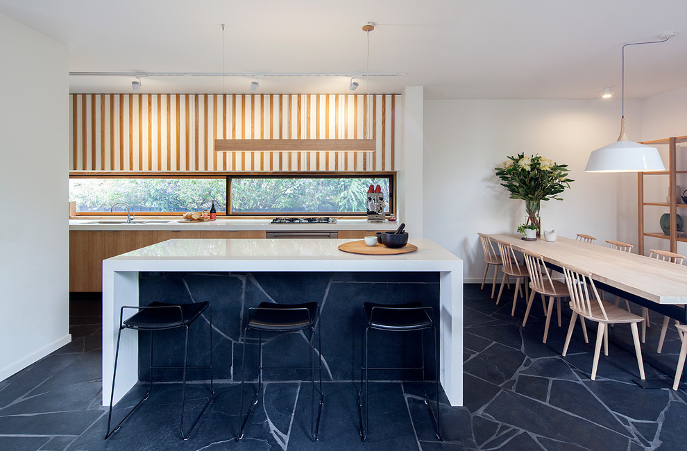 Exemple d'une cuisine américaine parallèle rétro en bois clair avec un évier encastré, un placard à porte plane, fenêtre, un électroménager en acier inoxydable, îlot, un sol noir et un plan de travail blanc.