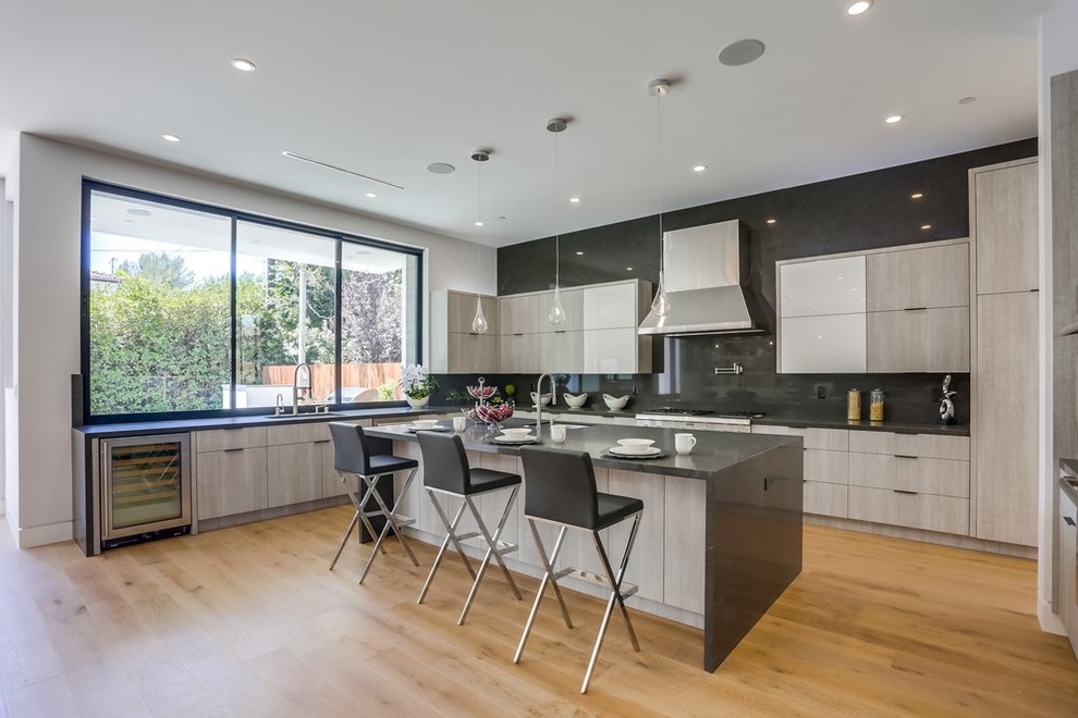 Inspiration for a large contemporary u-shaped open plan kitchen in Los Angeles with a triple-bowl sink, flat-panel cabinets, medium wood cabinets, composite countertops, grey splashback, stainless steel appliances, light hardwood flooring, an island, beige floors and grey worktops.
