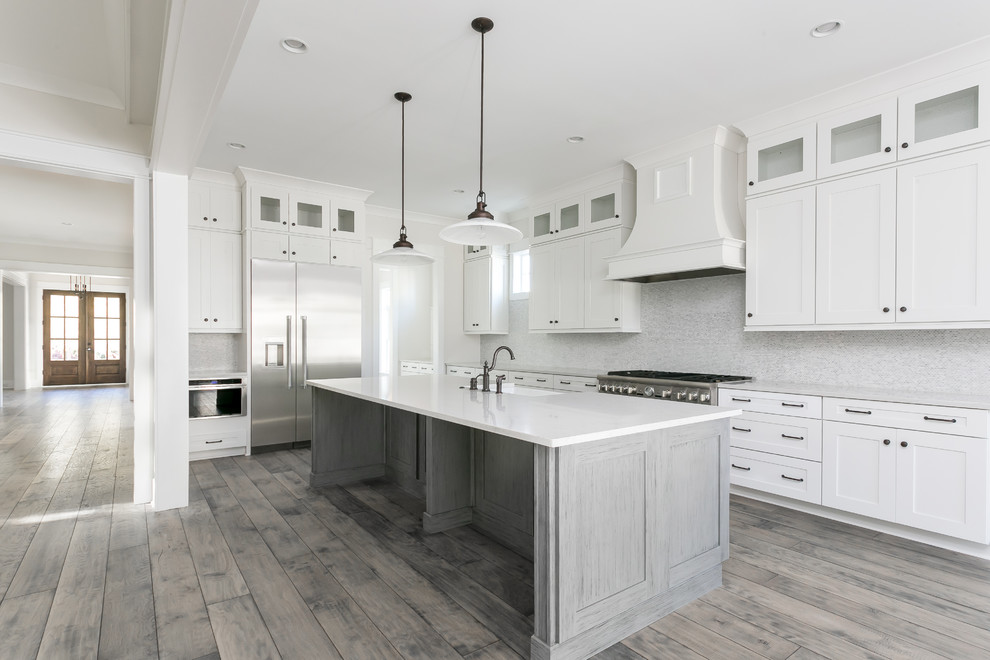 Lantlig inredning av ett stort vit vitt kök, med en rustik diskho, släta luckor, vita skåp, bänkskiva i kvarts, grått stänkskydd, stänkskydd i marmor, rostfria vitvaror, mellanmörkt trägolv, en köksö och grått golv