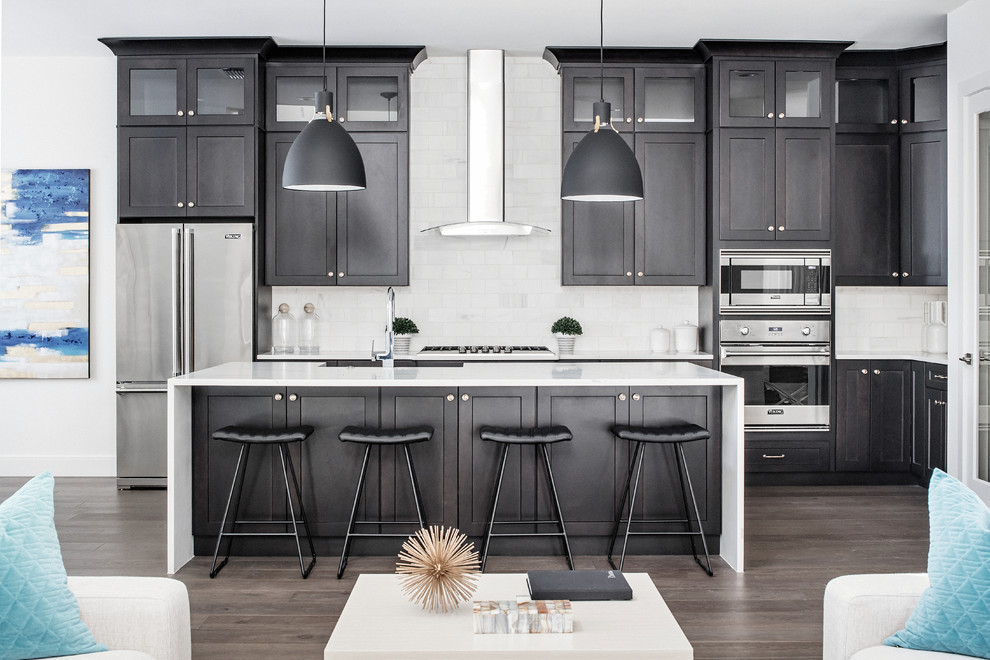 Traditional grey and white l-shaped open plan kitchen in Orlando with shaker cabinets, black cabinets, engineered stone countertops, white splashback, marble splashback, stainless steel appliances, dark hardwood flooring, an island and brown floors.