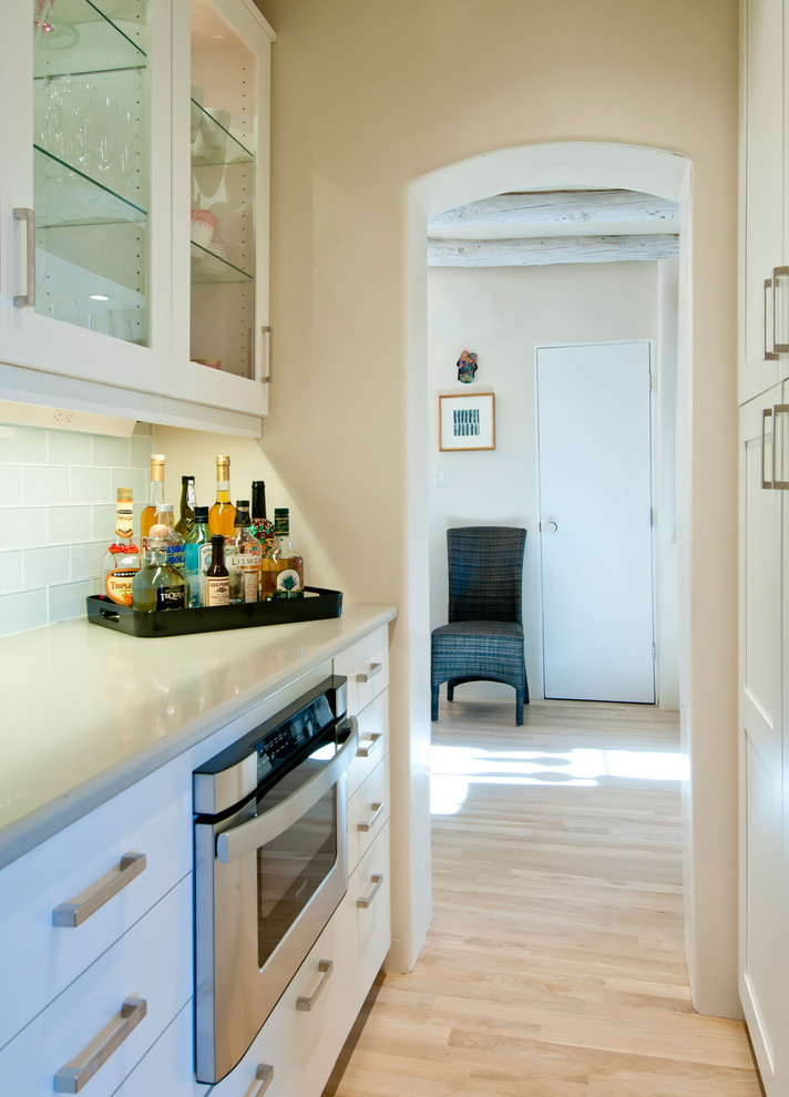 Amerikansk inredning av ett mellanstort l-kök, med en undermonterad diskho, skåp i shakerstil, vita skåp, blått stänkskydd, stänkskydd i glaskakel, rostfria vitvaror, ljust trägolv och beiget golv