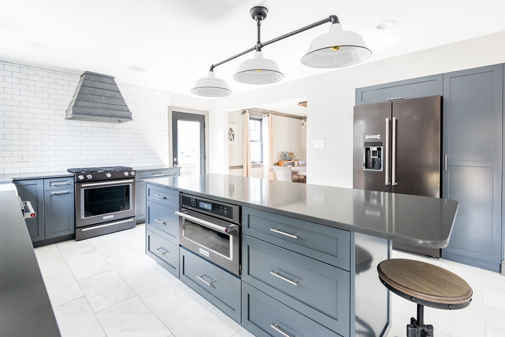 This is an example of a large farmhouse l-shaped enclosed kitchen in Wilmington with a belfast sink, shaker cabinets, white splashback, marble flooring, an island, blue cabinets, engineered stone countertops, metro tiled splashback, black appliances, grey floors and black worktops.