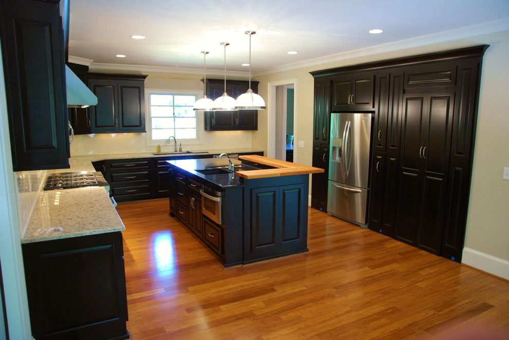Example of a mid-sized classic l-shaped medium tone wood floor and brown floor open concept kitchen design in Atlanta with an undermount sink, raised-panel cabinets, black cabinets, granite countertops, white backsplash, ceramic backsplash, stainless steel appliances and an island