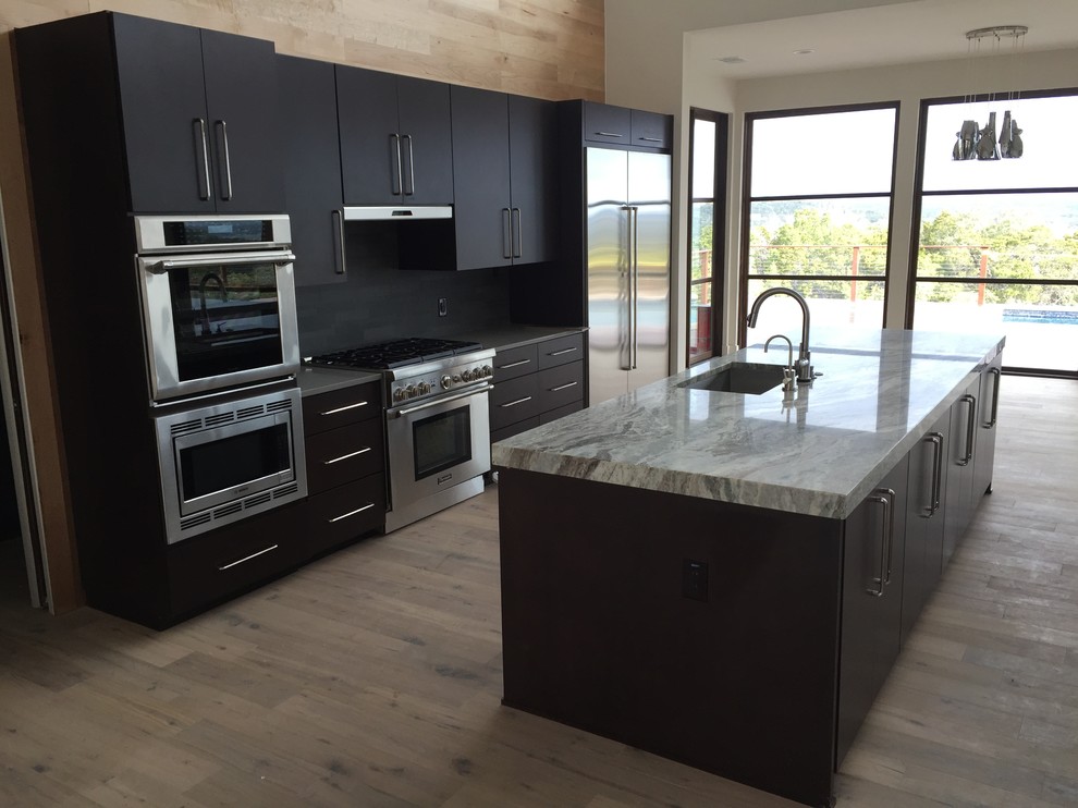 This is an example of a large contemporary galley open plan kitchen in Austin with a submerged sink, flat-panel cabinets, dark wood cabinets, granite worktops, black splashback, stone tiled splashback, stainless steel appliances, light hardwood flooring and multiple islands.