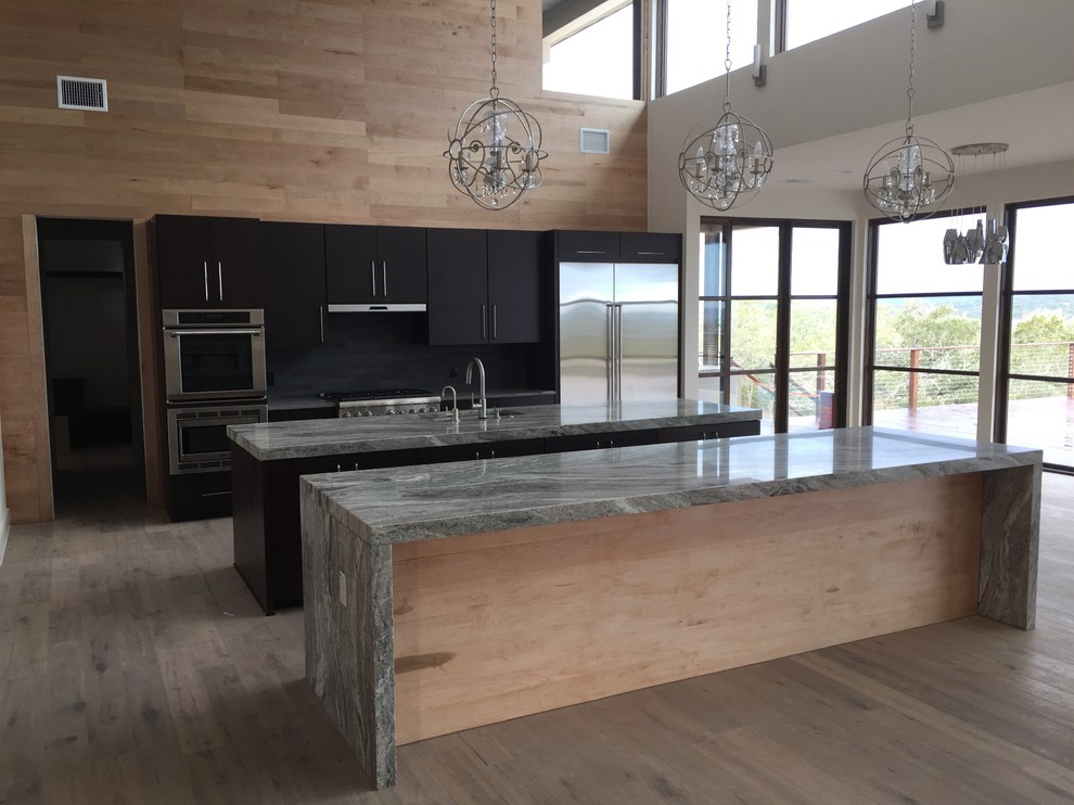 Photo of a large contemporary galley open plan kitchen in Austin with a submerged sink, flat-panel cabinets, dark wood cabinets, granite worktops, black splashback, stone tiled splashback, stainless steel appliances, light hardwood flooring and multiple islands.