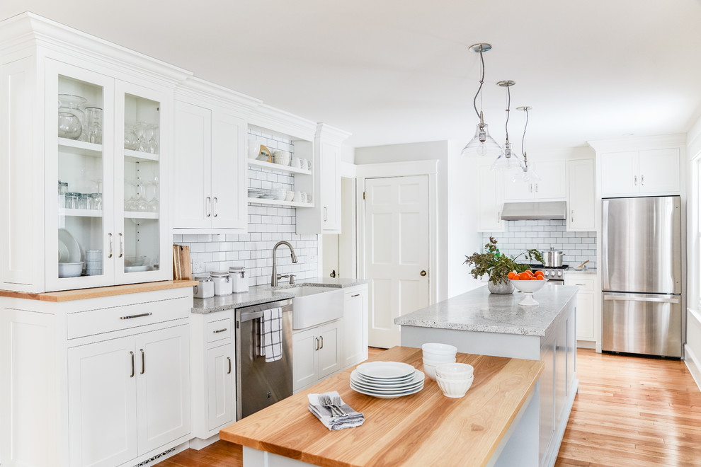 Classic l-shaped kitchen in Portland Maine with a belfast sink, shaker cabinets, white cabinets, white splashback, metro tiled splashback, stainless steel appliances, medium hardwood flooring, an island, brown floors and grey worktops.