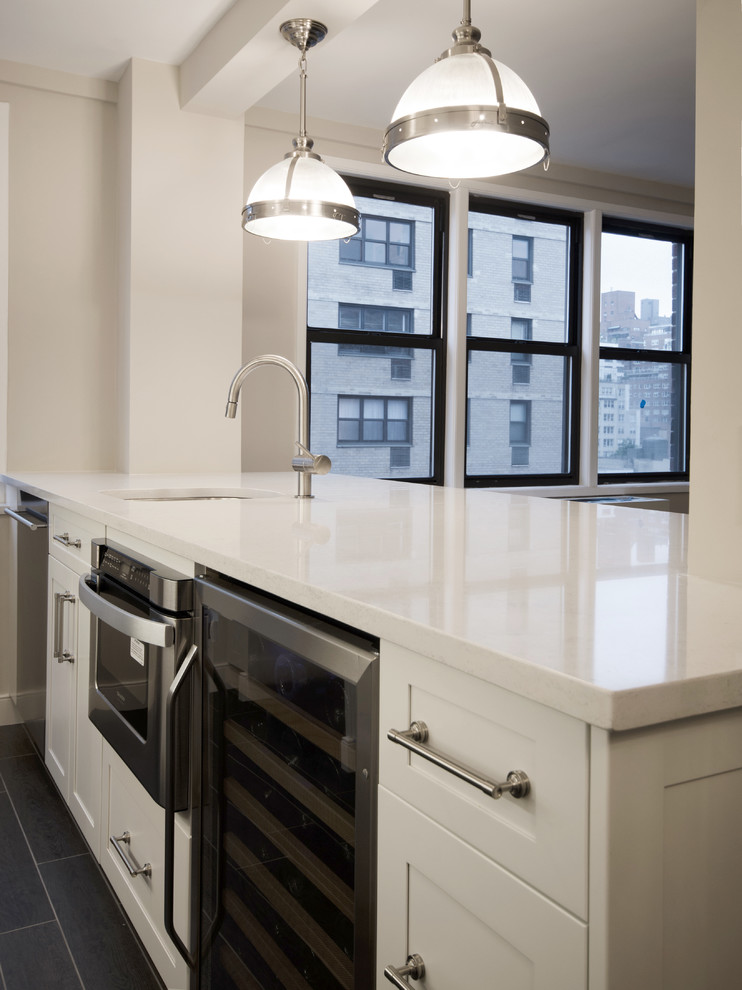 This is an example of a small modern galley kitchen/diner in New York with an island, shaker cabinets, white cabinets, quartz worktops, white splashback, stainless steel appliances, a submerged sink, porcelain flooring and glass tiled splashback.