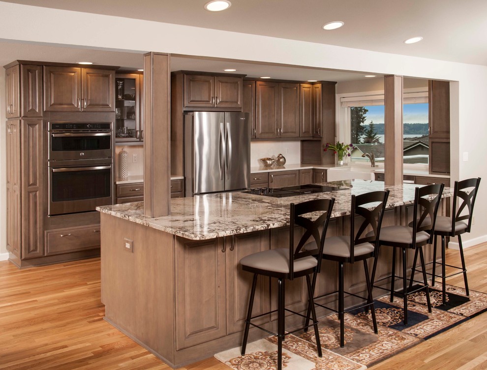 Inspiration for a traditional l-shaped kitchen in Seattle with a belfast sink, raised-panel cabinets, dark wood cabinets, beige splashback, stainless steel appliances, light hardwood flooring, an island, beige worktops and granite worktops.
