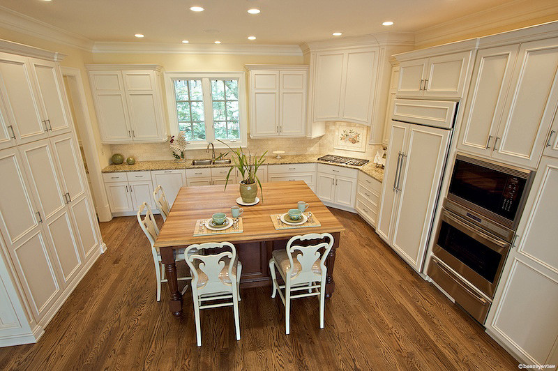 Open concept kitchen - large traditional l-shaped medium tone wood floor open concept kitchen idea in Atlanta with an undermount sink, recessed-panel cabinets, white cabinets, wood countertops, beige backsplash, stone tile backsplash, stainless steel appliances and an island