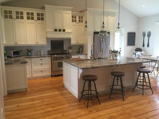 This is an example of a country l-shaped kitchen/diner in Toronto with a belfast sink, shaker cabinets, white cabinets, granite worktops, white splashback, ceramic splashback, stainless steel appliances, light hardwood flooring and an island.