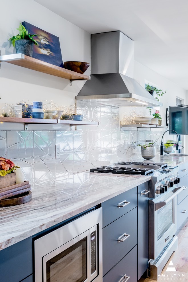Inredning av ett modernt stort vit vitt kök, med en rustik diskho, släta luckor, blå skåp, bänkskiva i kvartsit, vitt stänkskydd, stänkskydd i porslinskakel, rostfria vitvaror, ljust trägolv och en köksö