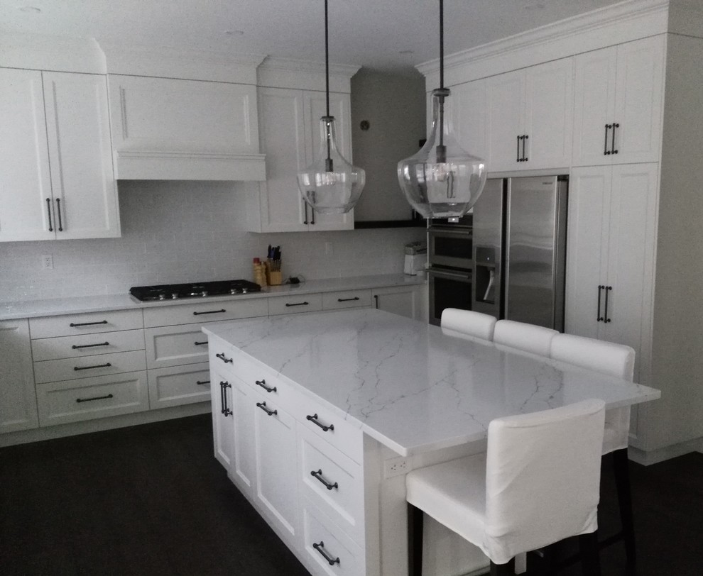 Inredning av ett klassiskt stort u-kök, med en undermonterad diskho, skåp i shakerstil, vita skåp, bänkskiva i kvartsit, vitt stänkskydd, integrerade vitvaror, mörkt trägolv, en köksö och brunt golv