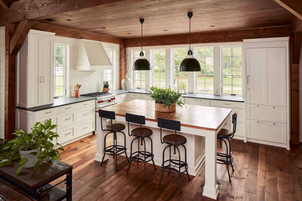Example of a cottage l-shaped dark wood floor and brown floor kitchen design in Milwaukee with a farmhouse sink, shaker cabinets, white cabinets, paneled appliances, an island and black countertops