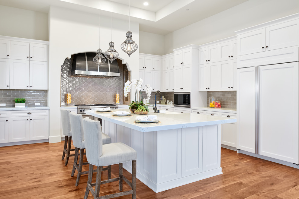 Inspiration for a large mediterranean l-shaped kitchen in Orange County with a belfast sink, white cabinets, engineered stone countertops, grey splashback, integrated appliances, medium hardwood flooring, an island, brown floors, white worktops, shaker cabinets and metro tiled splashback.