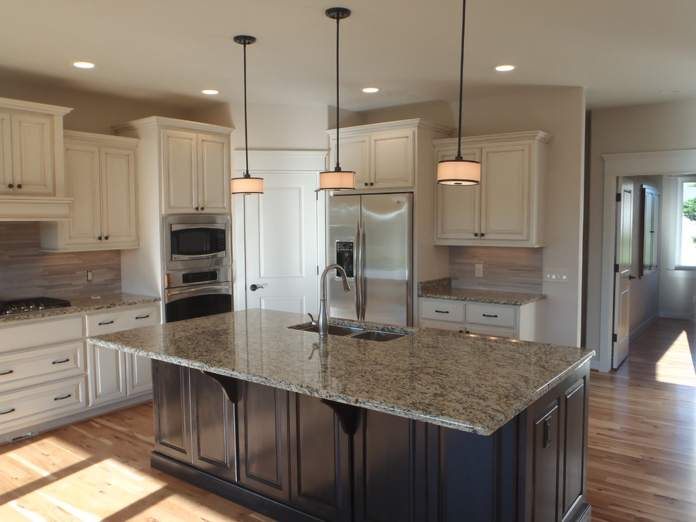 Inspiration for a timeless medium tone wood floor kitchen pantry remodel in Milwaukee with a double-bowl sink, raised-panel cabinets, distressed cabinets, granite countertops, gray backsplash, stone tile backsplash, stainless steel appliances and an island