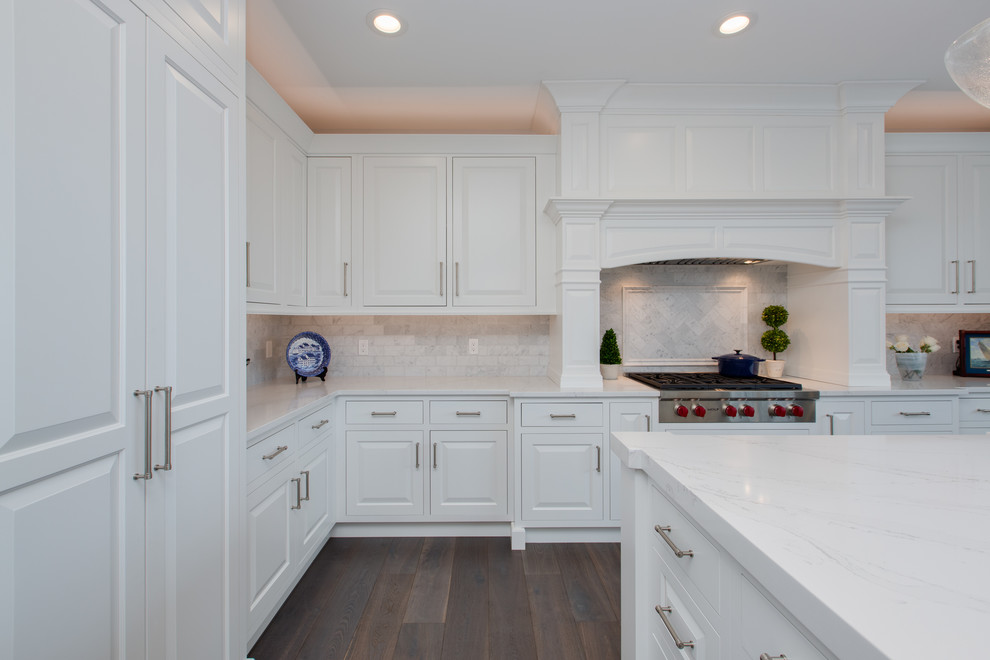 Klassisk inredning av ett stort vit vitt kök, med en undermonterad diskho, luckor med upphöjd panel, vita skåp, bänkskiva i kvarts, vitt stänkskydd, stänkskydd i tunnelbanekakel, rostfria vitvaror, mellanmörkt trägolv, en köksö och brunt golv