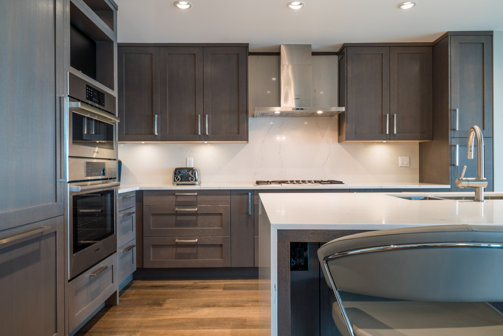 Moderne Wohnküche in L-Form mit Doppelwaschbecken, Schrankfronten im Shaker-Stil, Quarzit-Arbeitsplatte, Rückwand aus Stein, Küchengeräten aus Edelstahl, dunklem Holzboden, Kücheninsel und weißer Arbeitsplatte in Vancouver