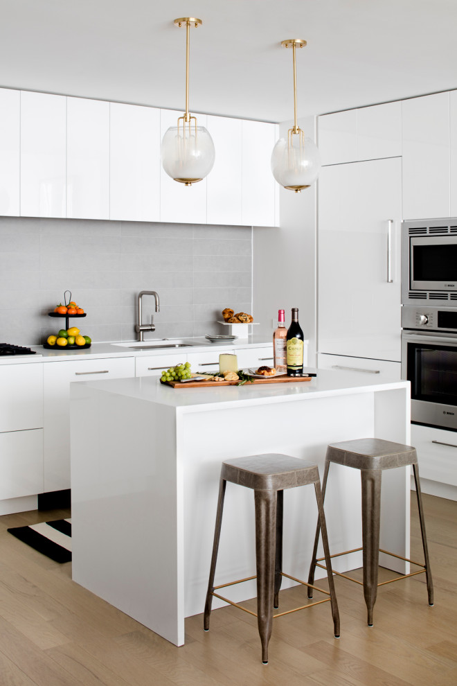 This is an example of a contemporary l-shaped kitchen in DC Metro with a submerged sink, flat-panel cabinets, white cabinets, grey splashback, stainless steel appliances, light hardwood flooring, an island, beige floors and white worktops.