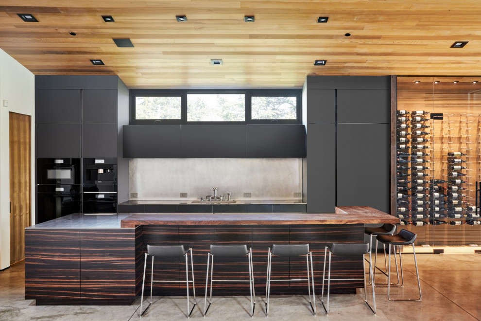 サンフランシスコにある広いコンテンポラリースタイルのおしゃれなキッチン (一体型シンク、フラットパネル扉のキャビネット、黒いキャビネット、ステンレスカウンター、黒い調理設備、コンクリートの床、グレーのキッチンパネル、木材のキッチンパネル、グレーの床、茶色いキッチンカウンター、板張り天井) の写真