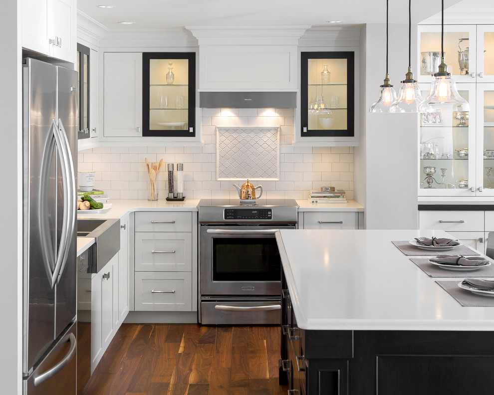 Traditional l-shaped kitchen in Vancouver with a belfast sink, glass-front cabinets, white splashback, metro tiled splashback, stainless steel appliances, medium hardwood flooring and an island.