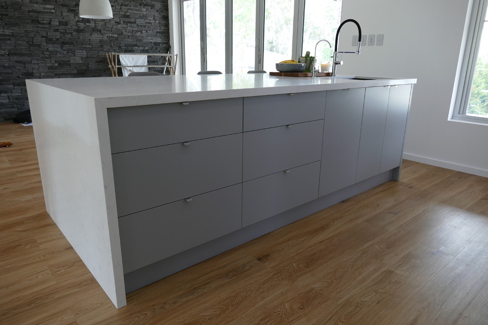 Photo of a medium sized modern galley open plan kitchen in Adelaide with a submerged sink, flat-panel cabinets, white cabinets, engineered stone countertops, white splashback, stone slab splashback, stainless steel appliances, medium hardwood flooring, an island, brown floors and white worktops.