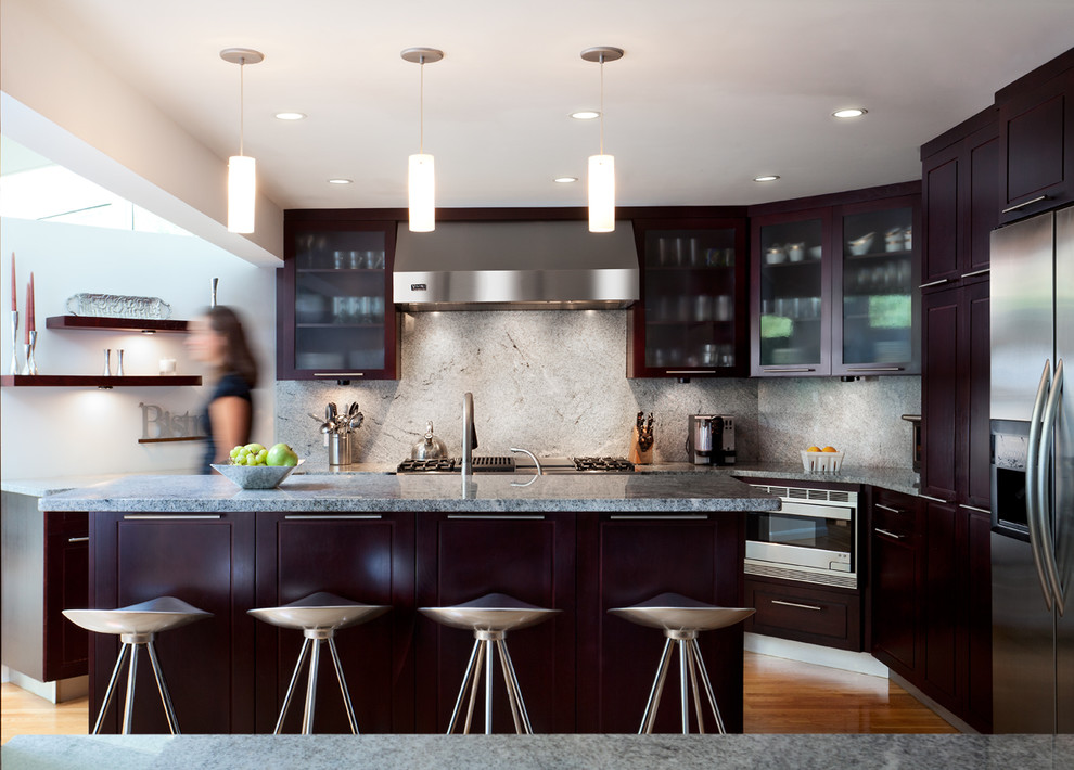 Modern inredning av ett kök, med luckor med glaspanel, skåp i mörkt trä, granitbänkskiva, grått stänkskydd, stänkskydd i sten, rostfria vitvaror, mellanmörkt trägolv och en köksö