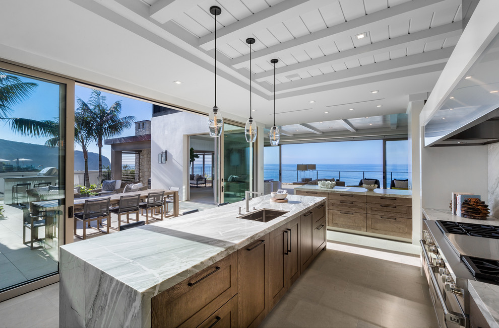 Inspiration for a coastal galley gray floor kitchen remodel in Orange County with an undermount sink, shaker cabinets, medium tone wood cabinets, white backsplash, stone slab backsplash, stainless steel appliances, an island and gray countertops