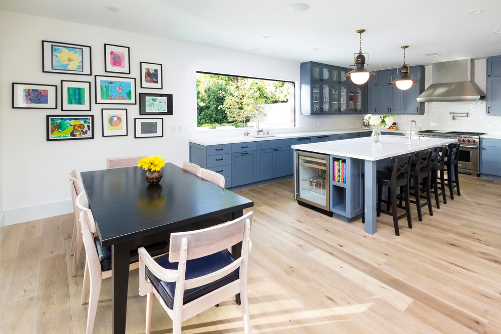 На фото: угловая кухня в стиле неоклассика (современная классика) с обеденным столом, врезной мойкой, фасадами в стиле шейкер, синими фасадами, белым фартуком и техникой из нержавеющей стали
