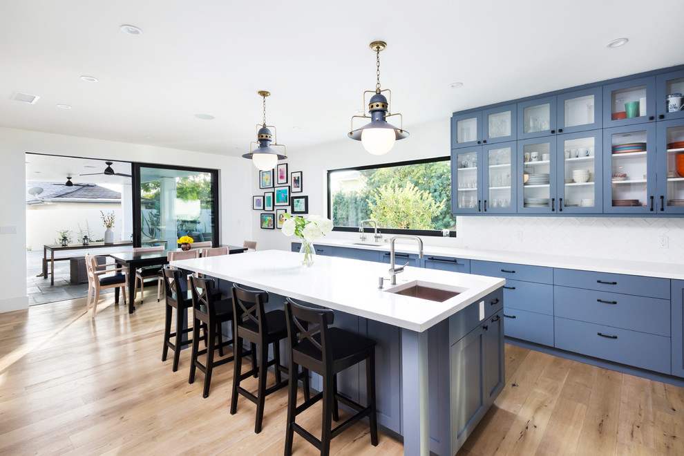 На фото: кухня в стиле неоклассика (современная классика) с обеденным столом, врезной мойкой, стеклянными фасадами, синими фасадами, белым фартуком и мойкой у окна